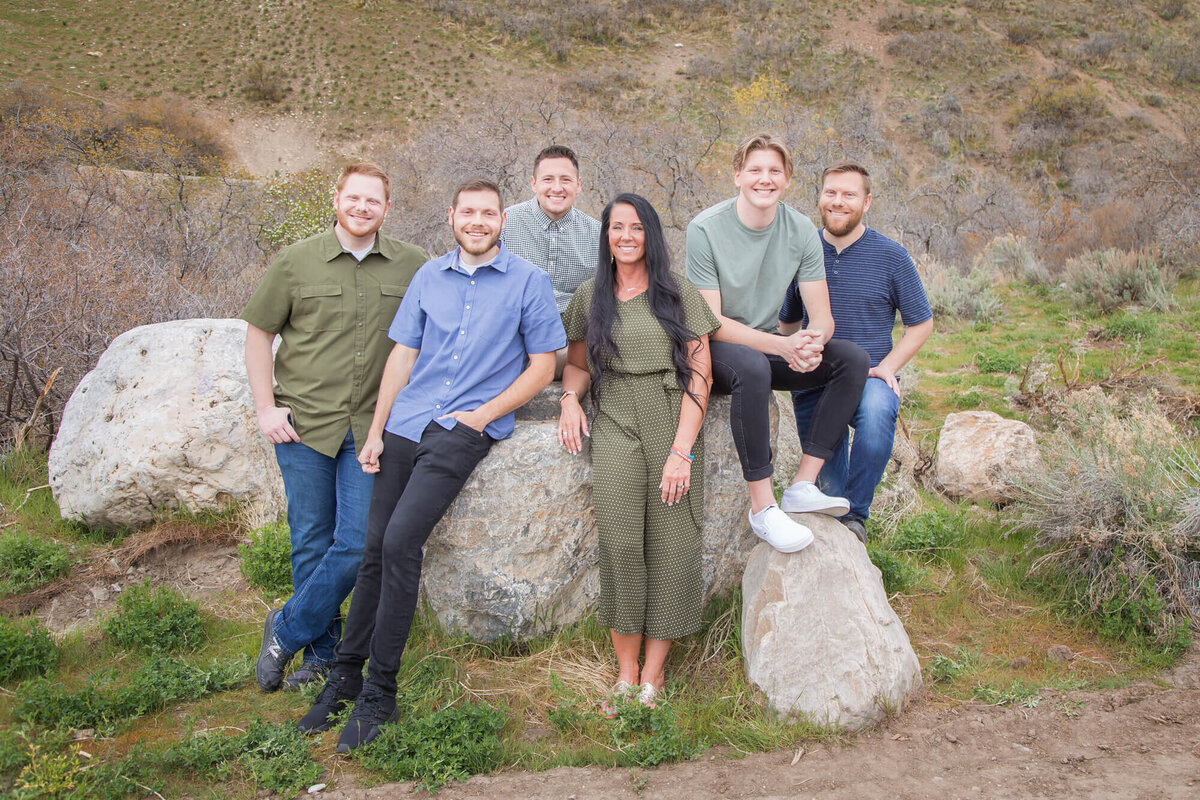 Drak brown haired woman and five adults sons in blue and green shirts sitting on a rock in Utah mountain park