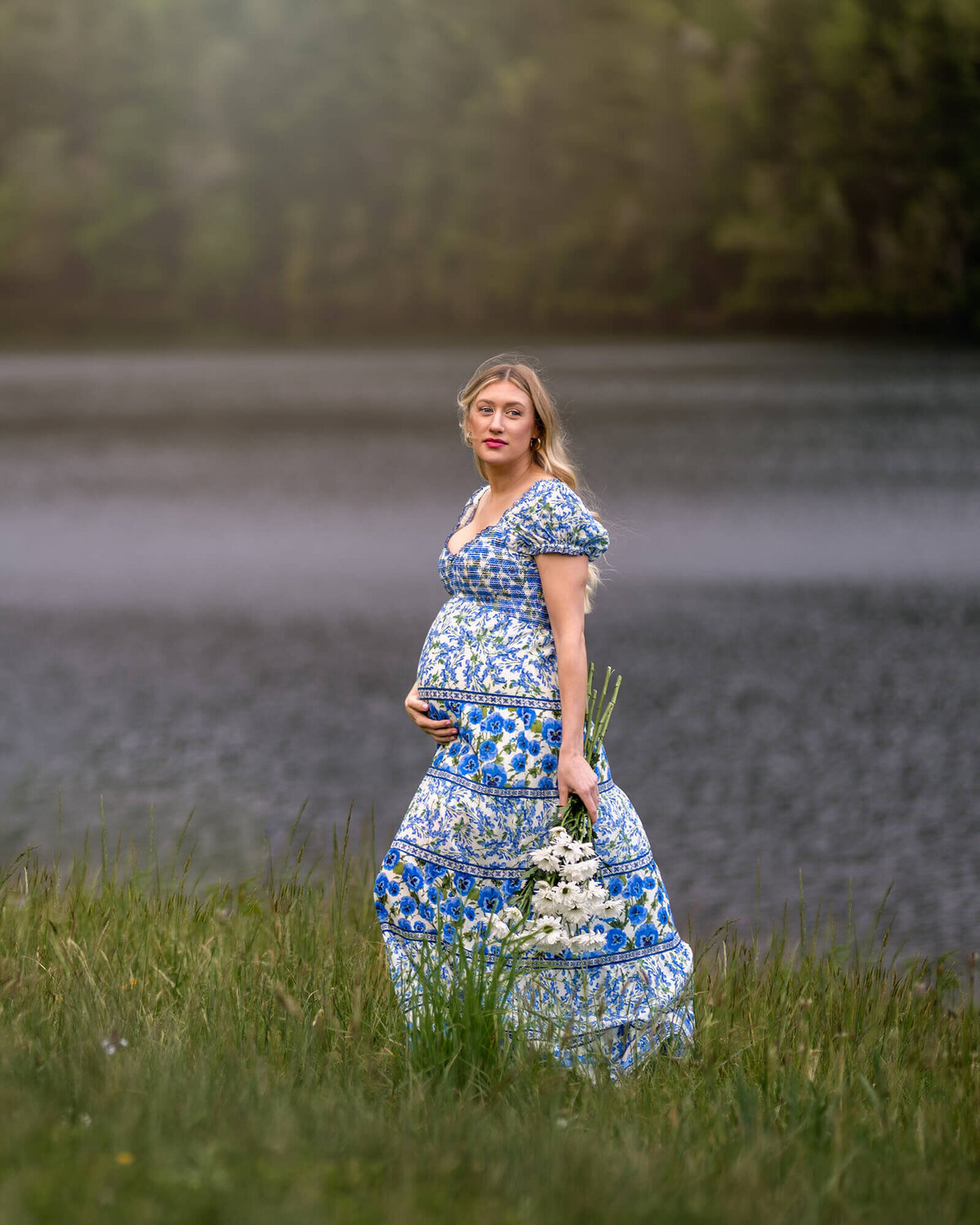 A gorgeous, young mama to be cradles her bump and holds flowers while standing in the grass by Fairfield Lake near Asheville