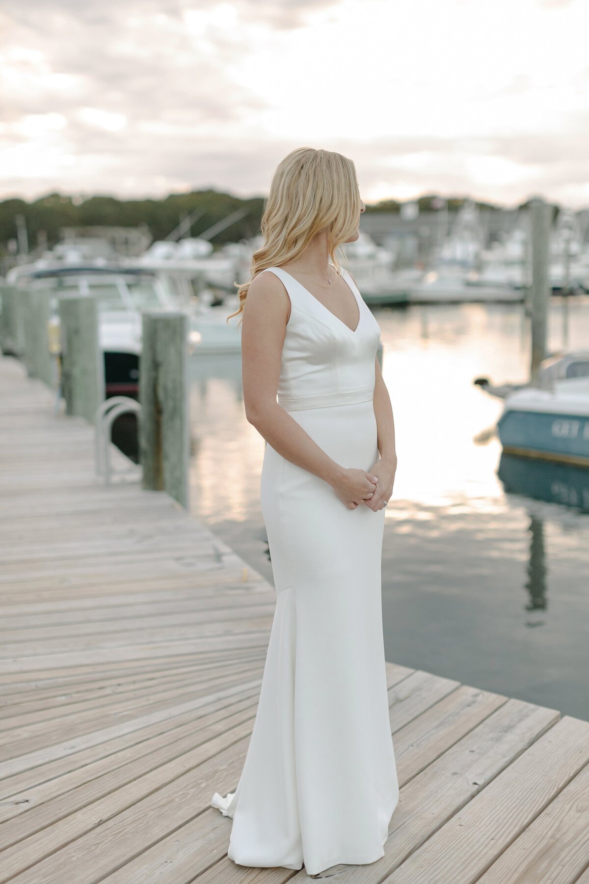 Bride stands on dock overlooking harbor at sunset.