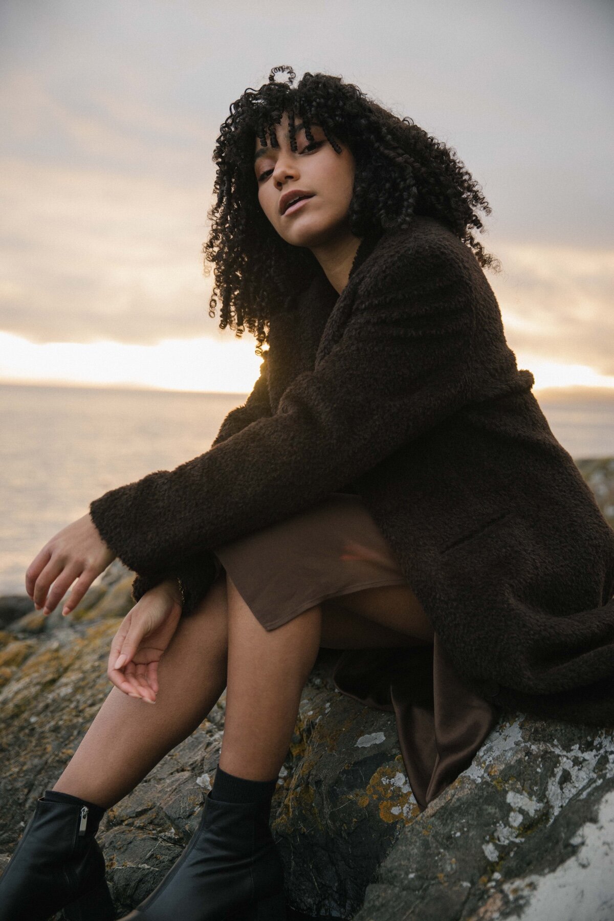 Picture of girl sitting on the rock at sunset in brown coat