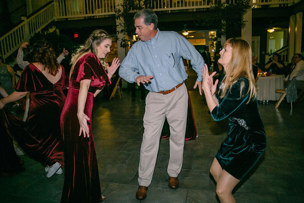 Three people, two women in velvet dresses and a man in a blue shirt and khakis, dance energetically together on a stone floor with a staircase and guests in the background.