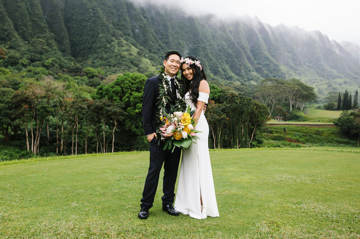 A bride and groom stand in front of a mountain range in Hawaii on their wedding day.