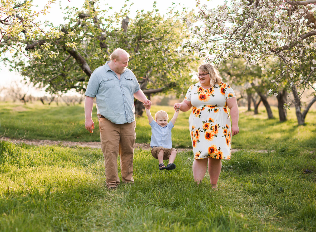 A family enjoying a sunny day in an orchard, with the parents lifting their child between them