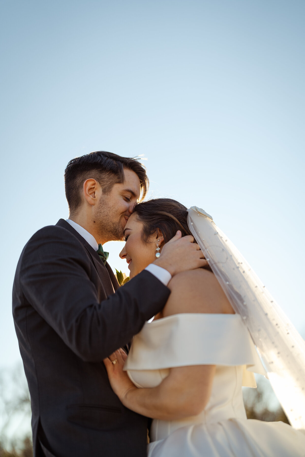 Moment captured as a groom tenderly kisses his bride's forehead during sunset at their DFW wedding. The bride's elegant veil flows softly as the couple embraces, highlighting the sophistication and intimacy of their wedding day.