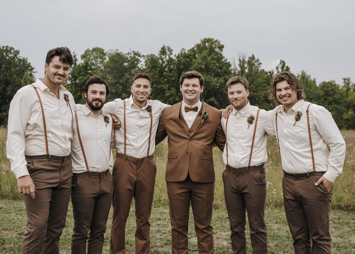 A group of six smiling groomsmen and a groom dressed in coordinated attire with suspenders and bow ties outdoors. Taken by jen Jarmuzek photography a Minneapolis wedding photographer