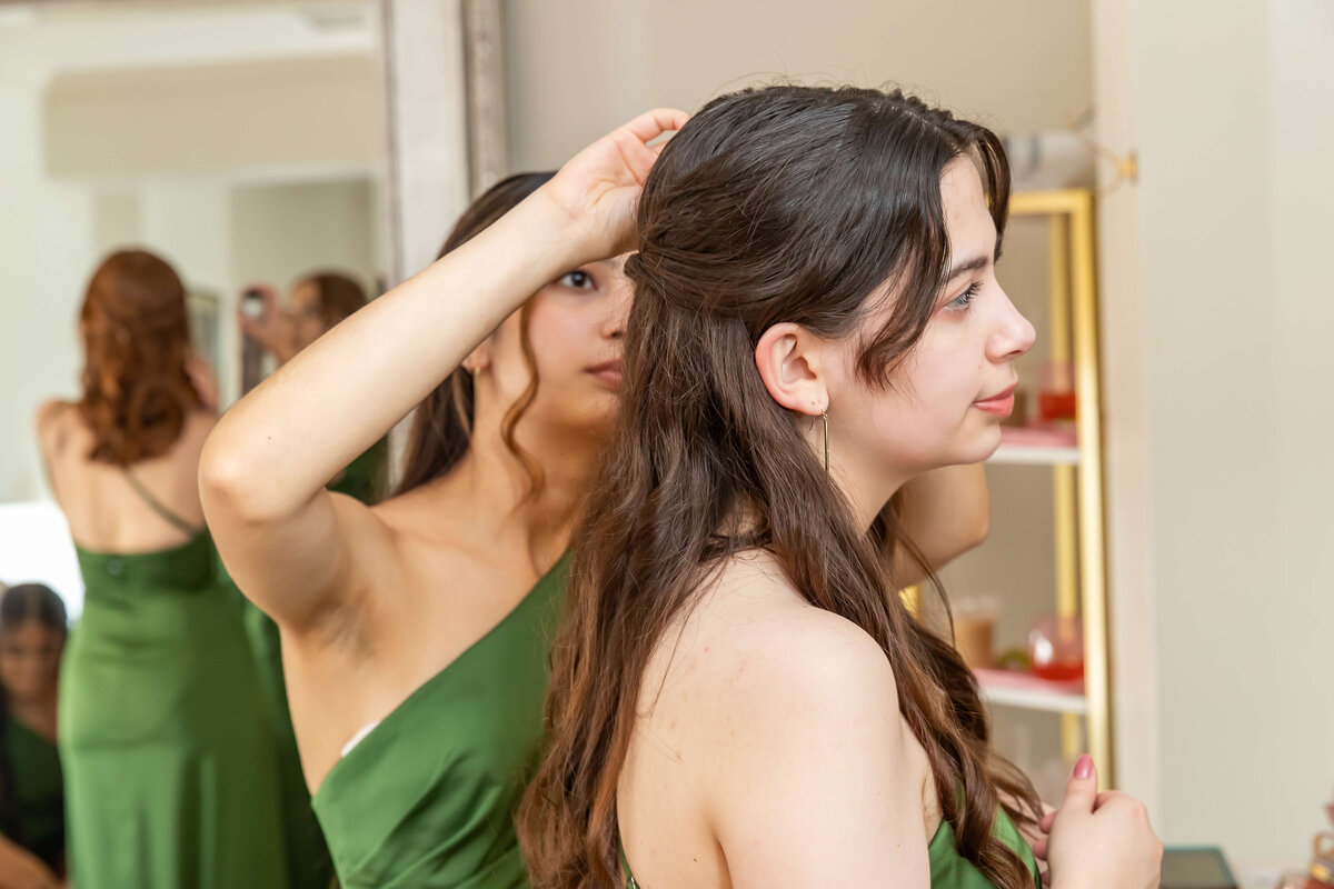 Two women wearing green dresses stand together indoors; one adjusts the others hair while they both face a mirror. Shelves and a reflection of one woman are visible in the background.