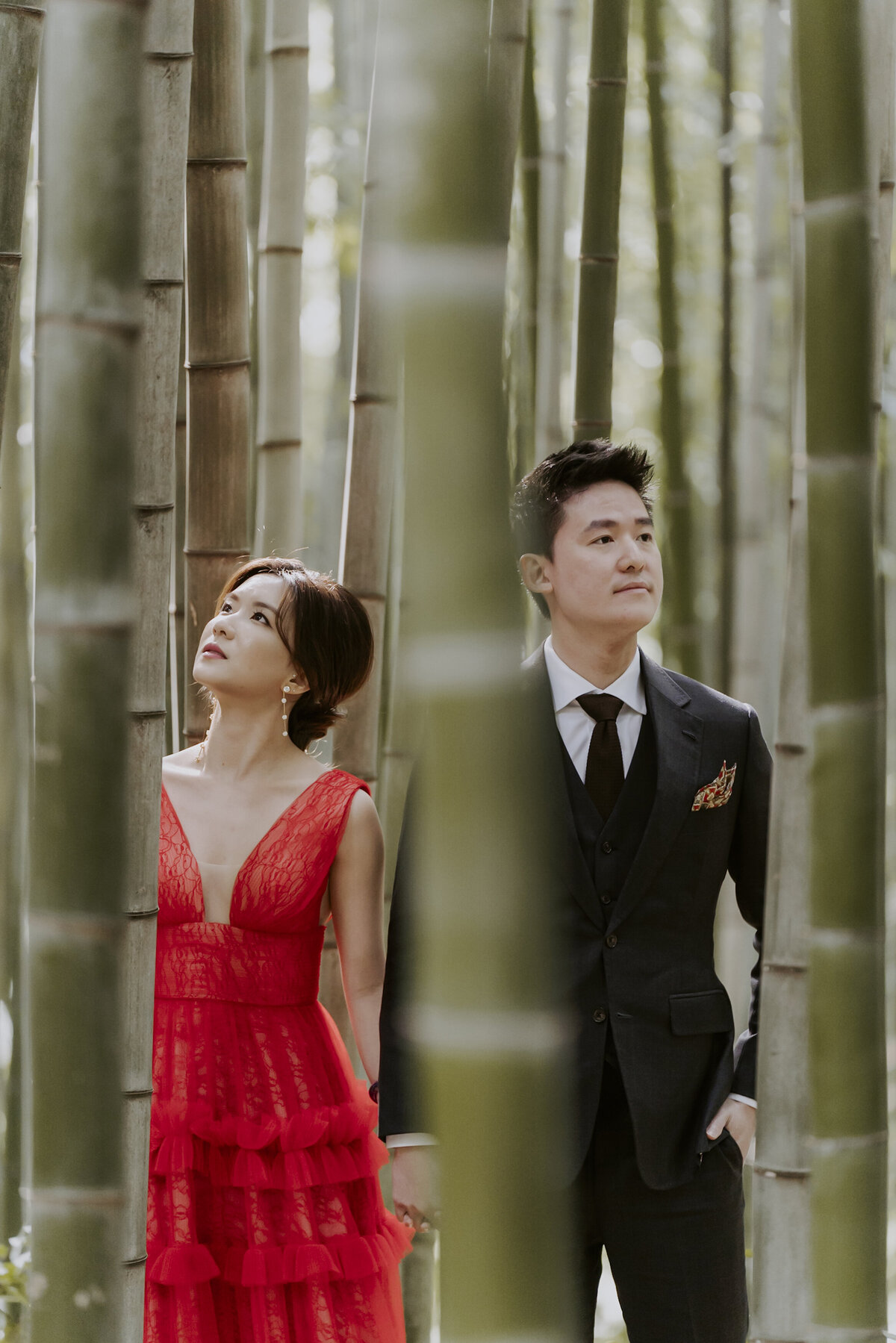 a woman wearing a red wedding dress and a man wearing a black suit look between the bamboo