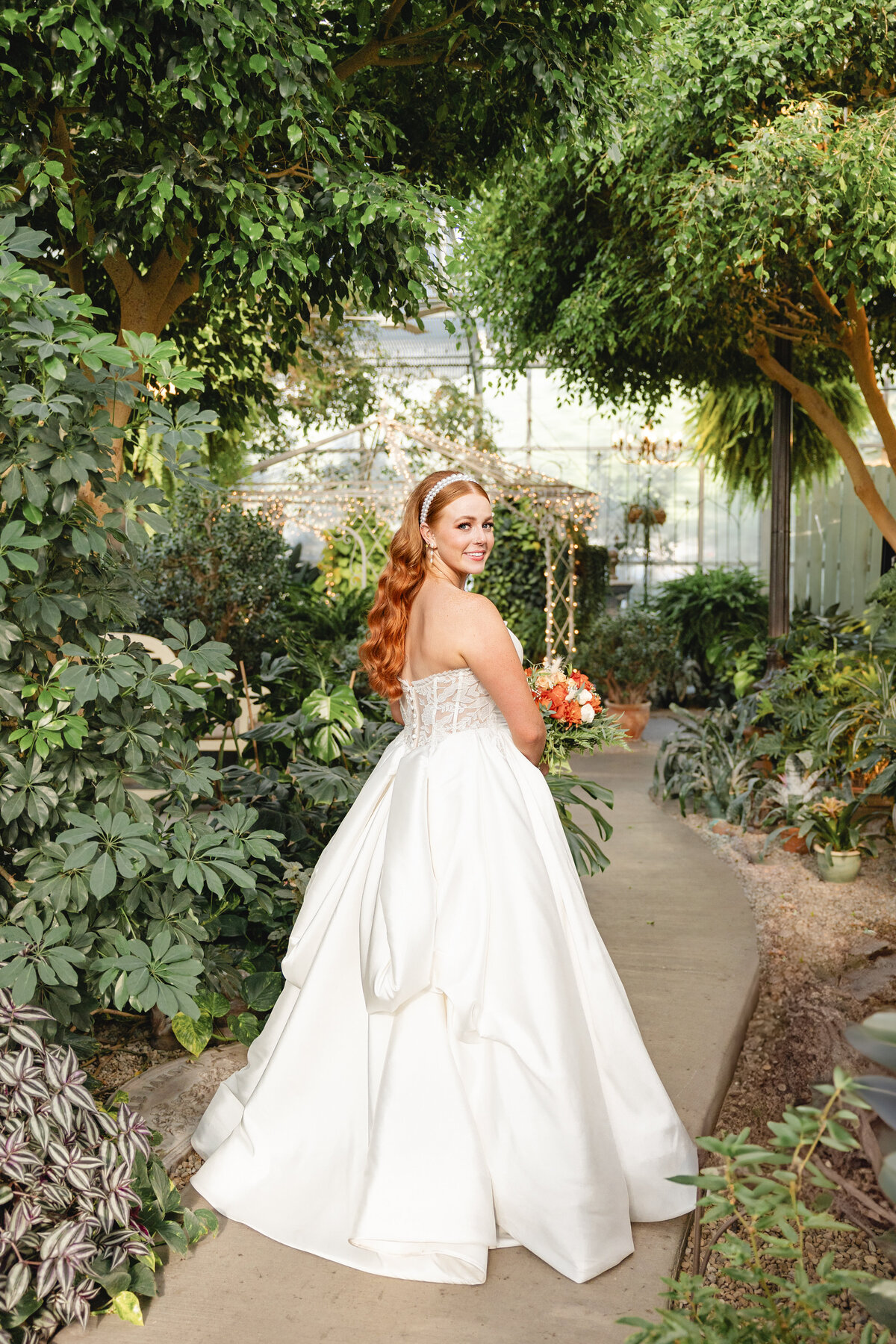 A bride with long red hair walks down a garden pathway at Le Jardin in Sandy, Utah, looking over her shoulder with a soft smile, holding a bouquet of orange and white flowers.
