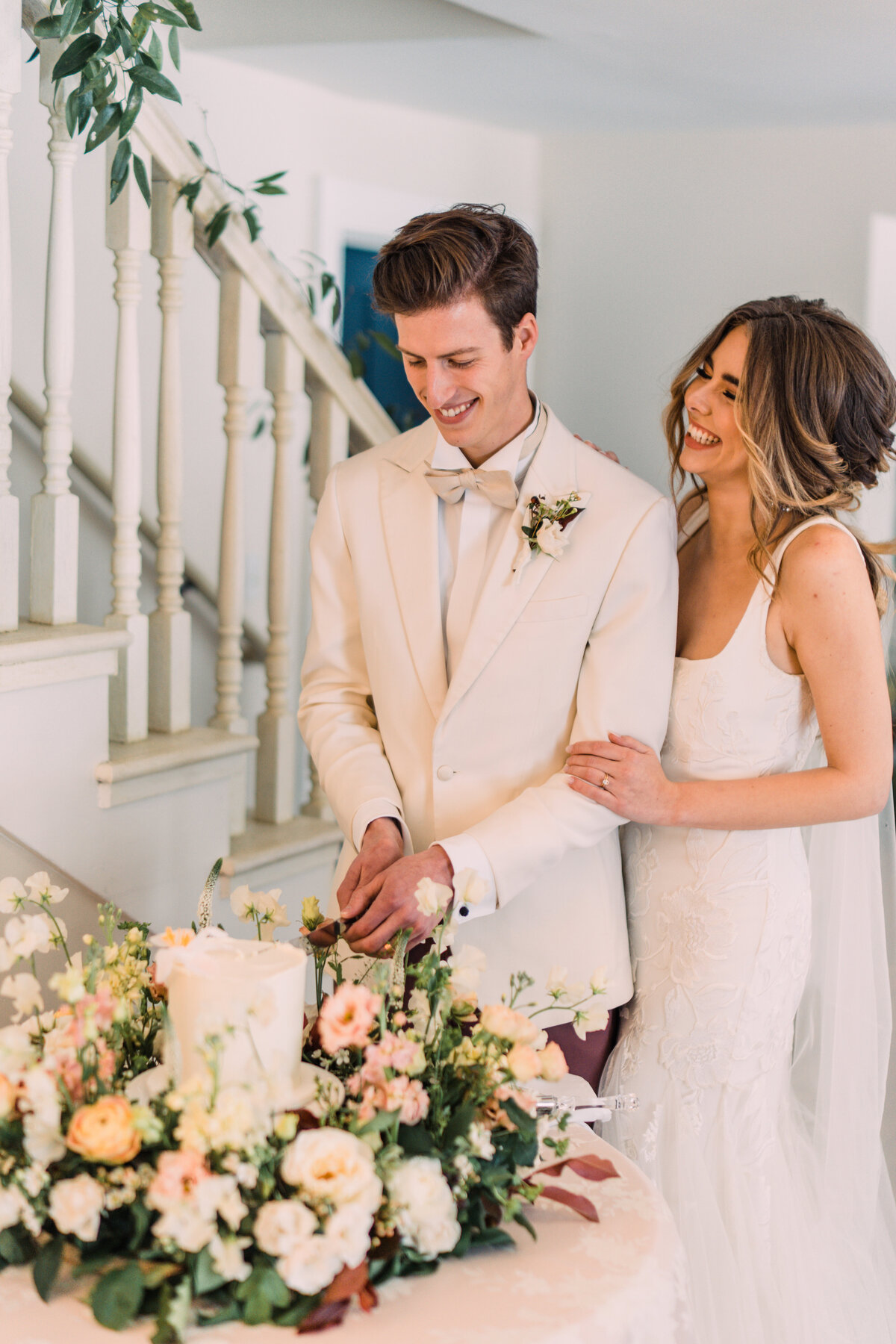 Bride laughing & holding groom's arm while he cuts a cake surrounded by a wreath of flowers at Zingerman's Cornman Farms