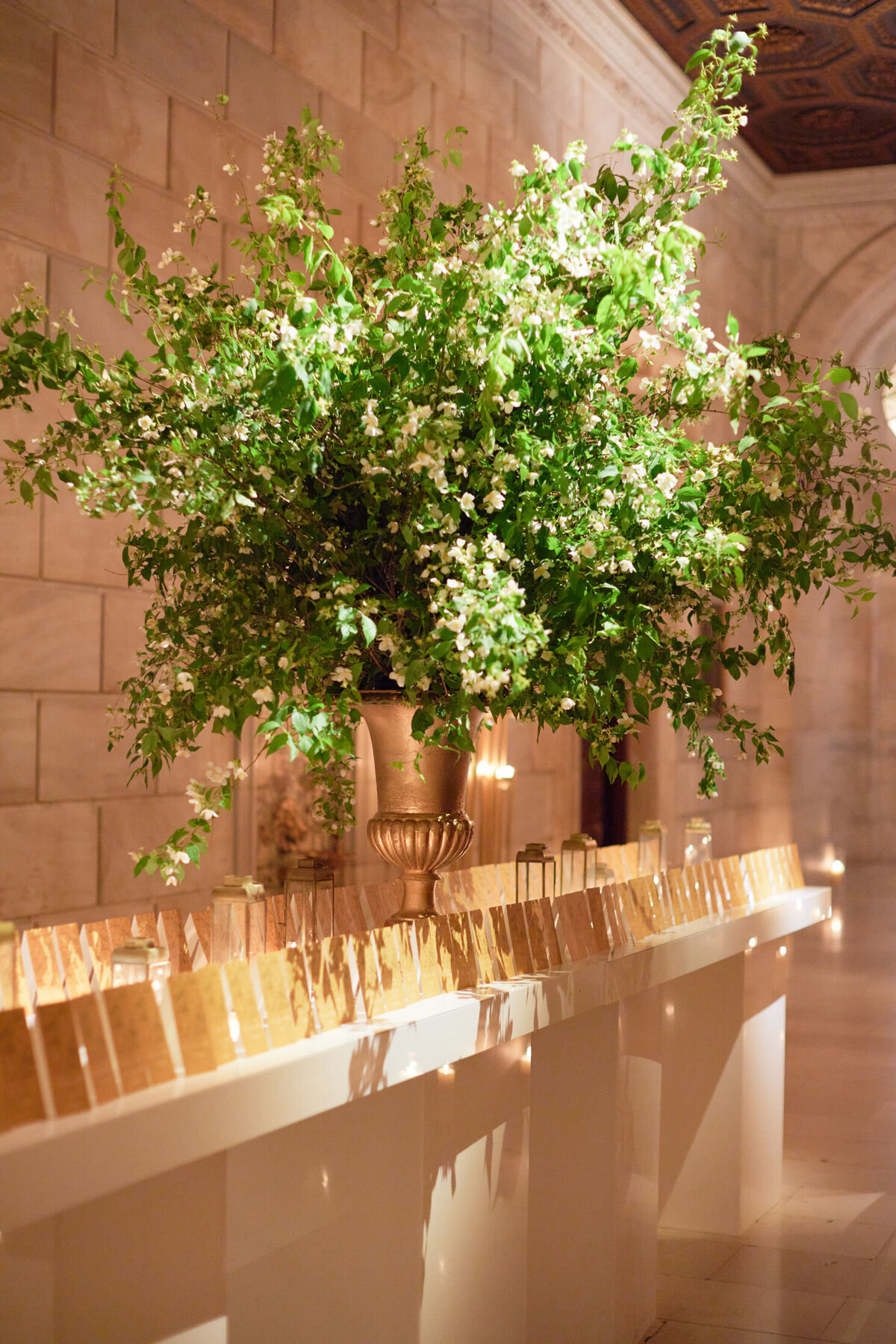 This elegant tablescape was captured at the iconic New York Public Library in New York City by the skilled photographer Shawn Connell with videography by Aaron Navak Films. The scene showcases a long table, beautifully decorated with numerous gold place cards, each delicately held by a small silver stand. The grand hall, with its impressive stone walls, provides a stunning backdrop for the setting. At the center of the table, a luxurious arrangement of green foliage and white flowers in a striking gold vase adds a touch of sophistication and charm to the overall ambiance.