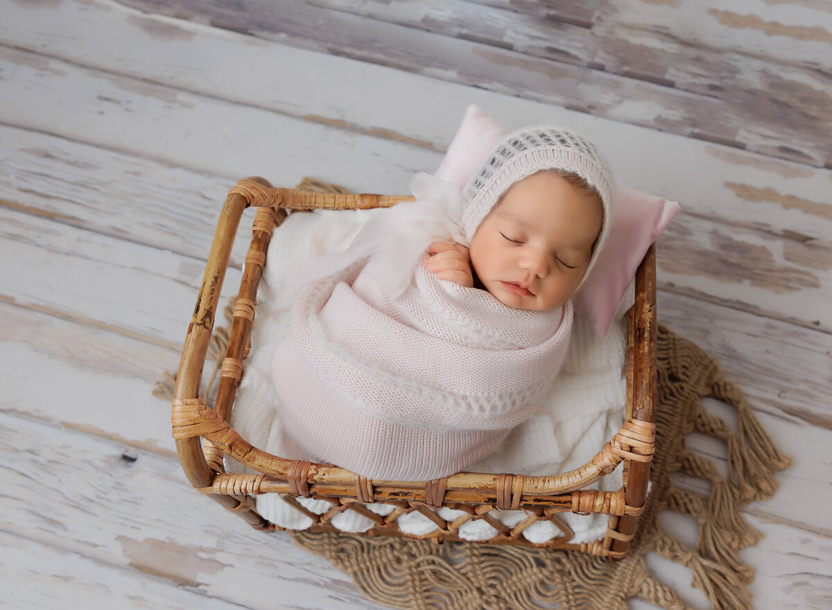 Baby girl in a pink swaddle sleeps in a basket for her brooklyn newborn photoshoot. Captured by top Brooklyn newborn photographer Chaya Bornstein