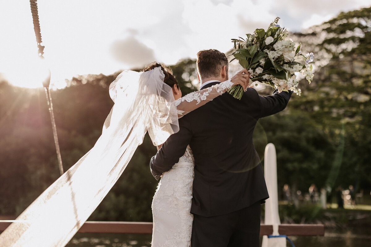 Bride and groom cheering turned towards guests on the shore from the boat