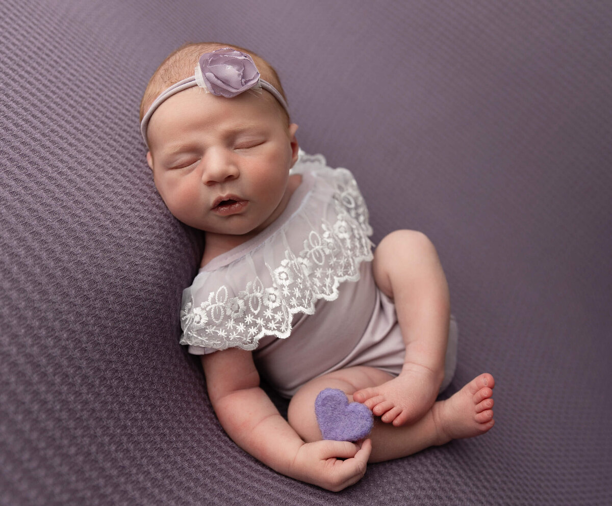 Newborn in a purple romper holding a purple heart in an Erie Pa photography studio in the Huck Finn pose