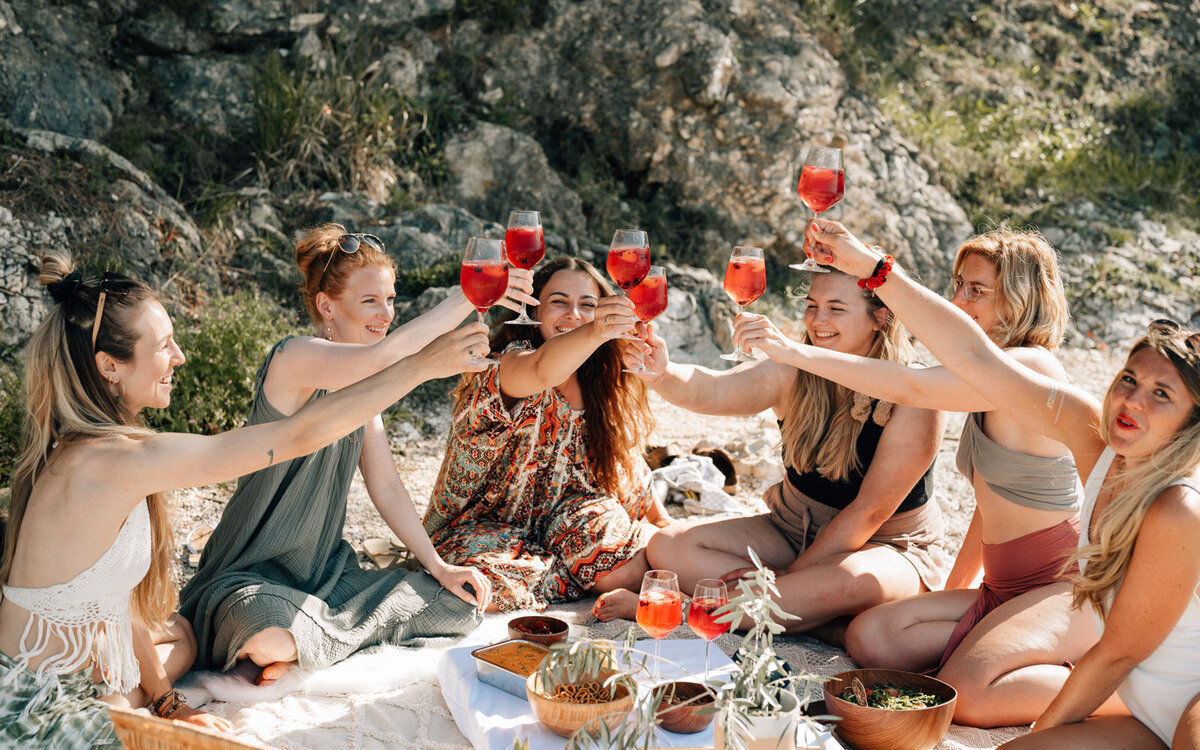 Frauen stoßen mit ihrem ayurvedischen Drink an während sie bei einem Picknick am Strand sitzen