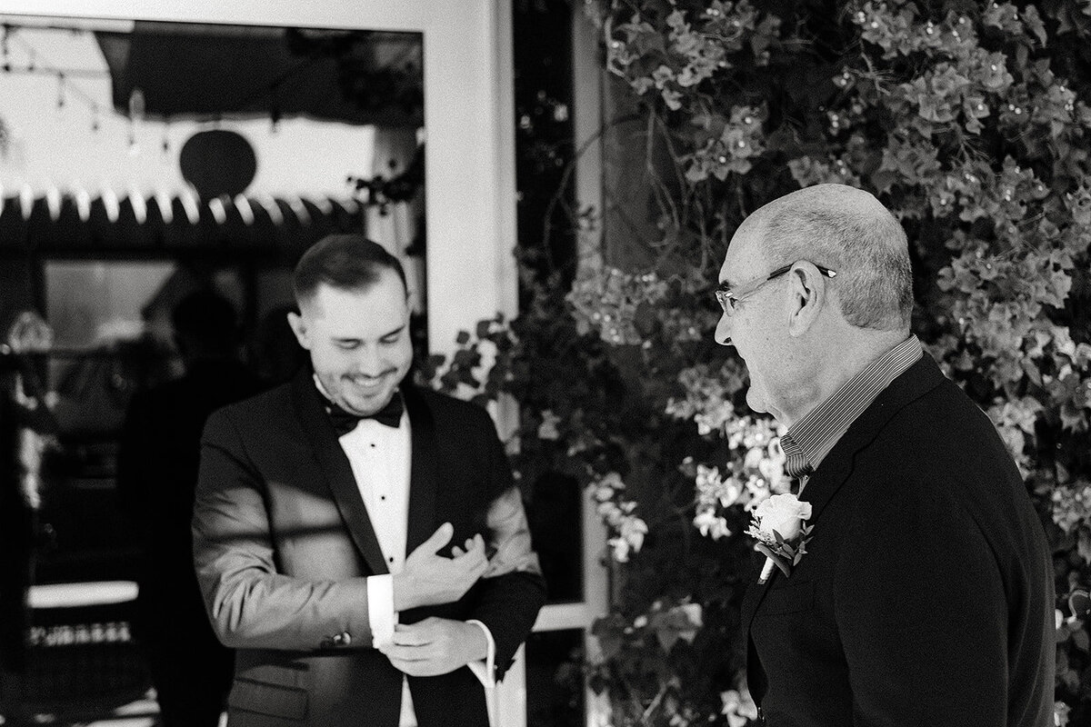 A candid black-and-white photo of the groom and his father sharing a laugh outdoors, with floral decor in the background.