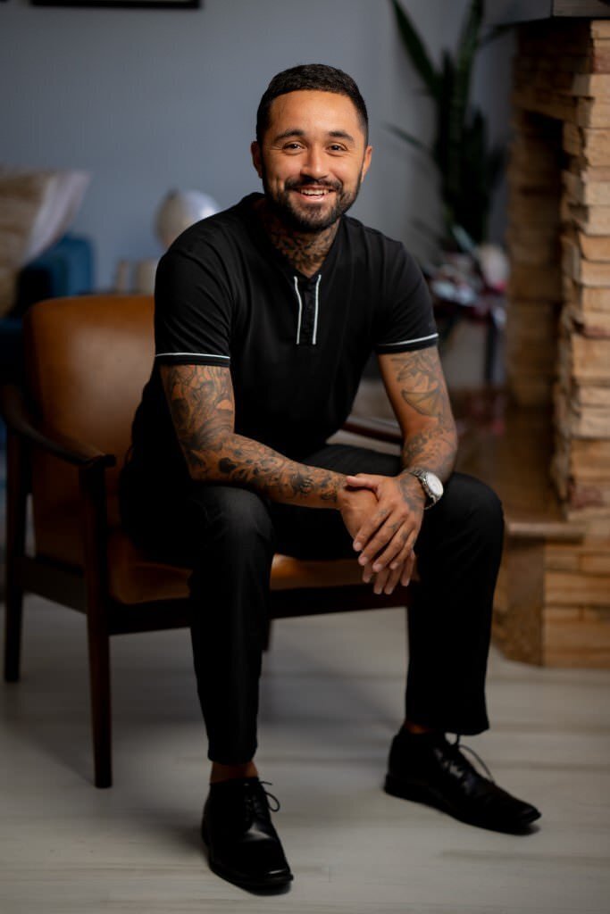 Smiling-man-with-tattooed-arms-sitting-on-a-chair-dressed-in-black