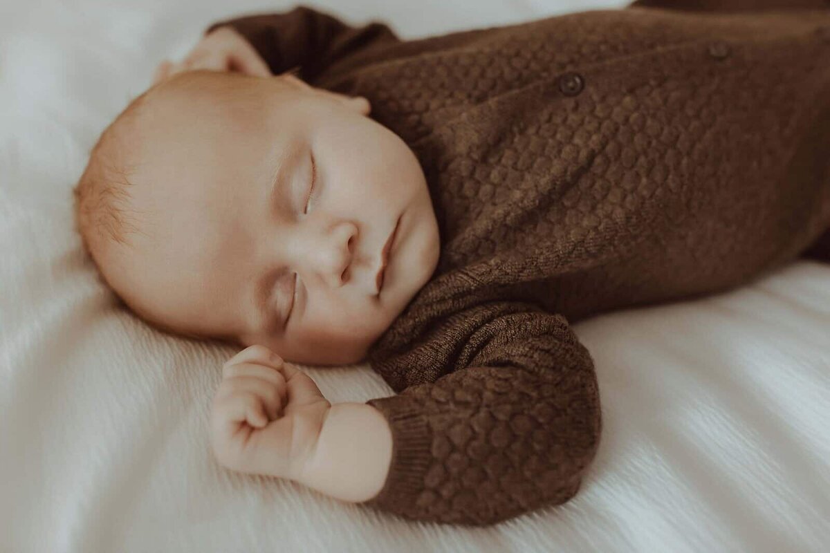 A baby sleeps on a white blanket during a Macon newborn photo session, wearing a textured brown outfit with buttons, one arm bent and eyes closed.