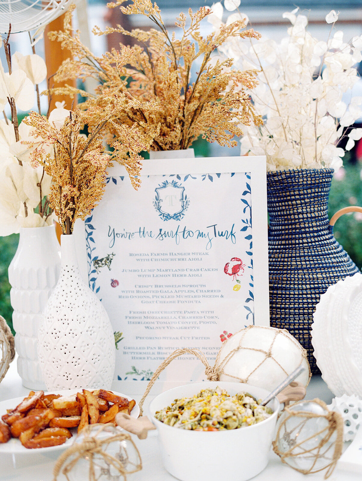 The picturesque setting for this event was The Hamptons, New York, at a stunning waterfront estate tented wedding captured beautifully by photographer Bonnie Sen and videographers East West Films. A table at the venue was elegantly decorated with a collection of decorative vases filled with dried flowers, complemented by a menu displayed in a charming blue and white frame. The menu featured a delightful message that read, You're the sun to my surf. The table arrangement also included an assortment of delectable dishes such as pretzels and a bowl of pasta salad, enhanced with decorative rope-covered buoys adding to the nautical theme.