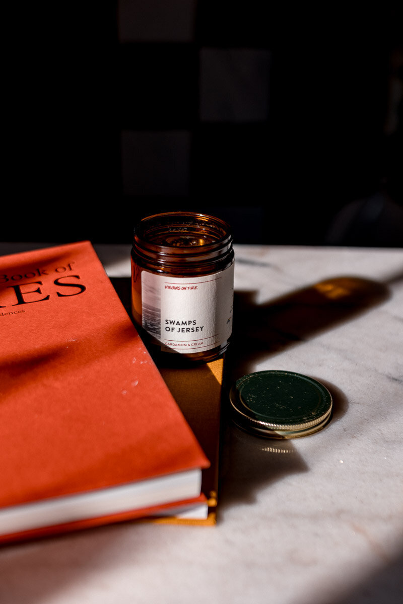 Virgins On Fire brand candle in scent "Jersey Swamps" on coffee table next to orange book