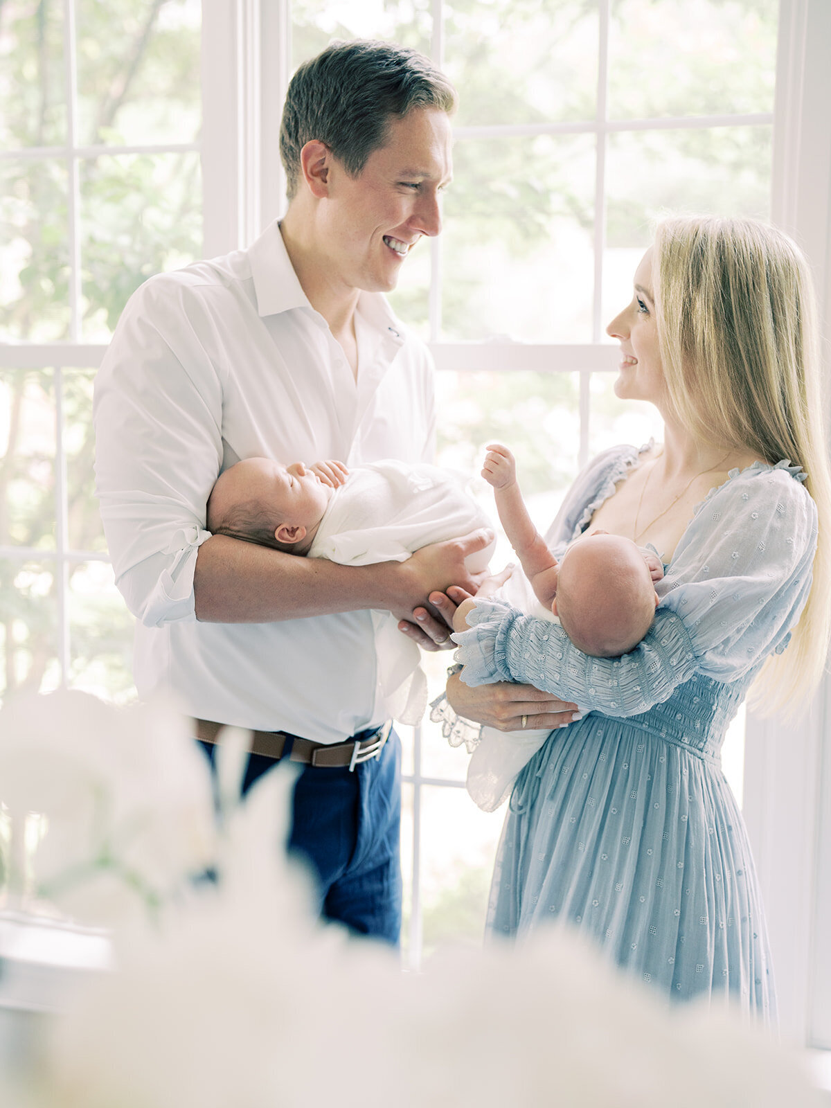 Blonde mother and father stand by a window holding their newborn twin boys smiling at one another during their Maryland newborn session.