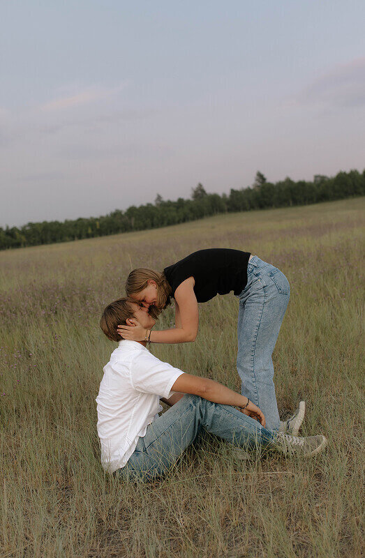 A man sits on the ground as his girlfriend is standing next to him. They're touching foreheads and she's holding his face.
