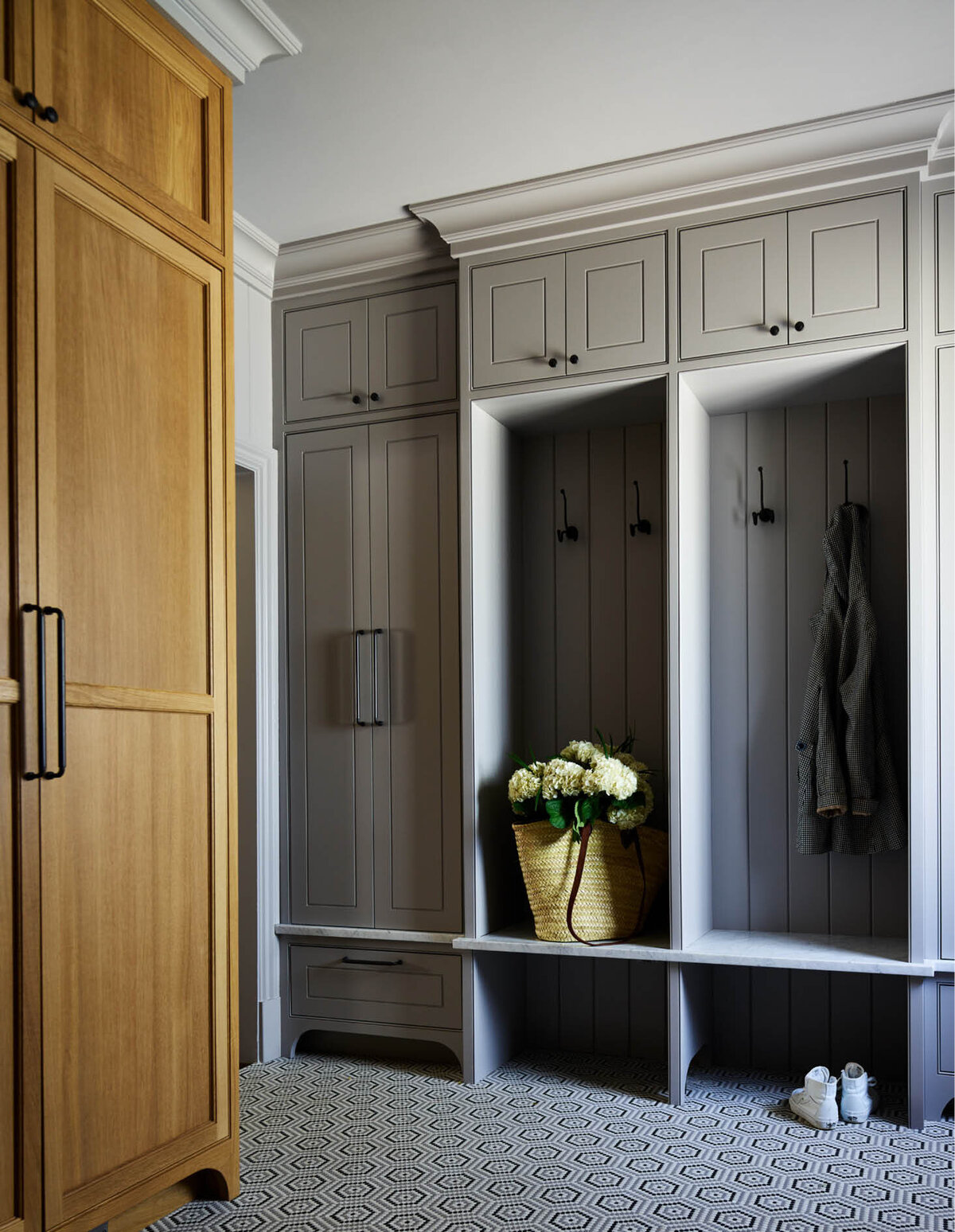 Next to tall oak cabinets, grey painted open and closed cabinets offer storage for coats, bags, shoes, and more, ensuring a functional and elegant mudroom.