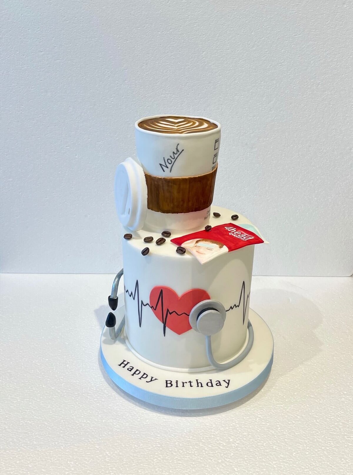 A medical themed two tier cake where the bottom tier shows a heart beat and stethoscope and the top tier is a cup of coffee with spilt coffee beans
