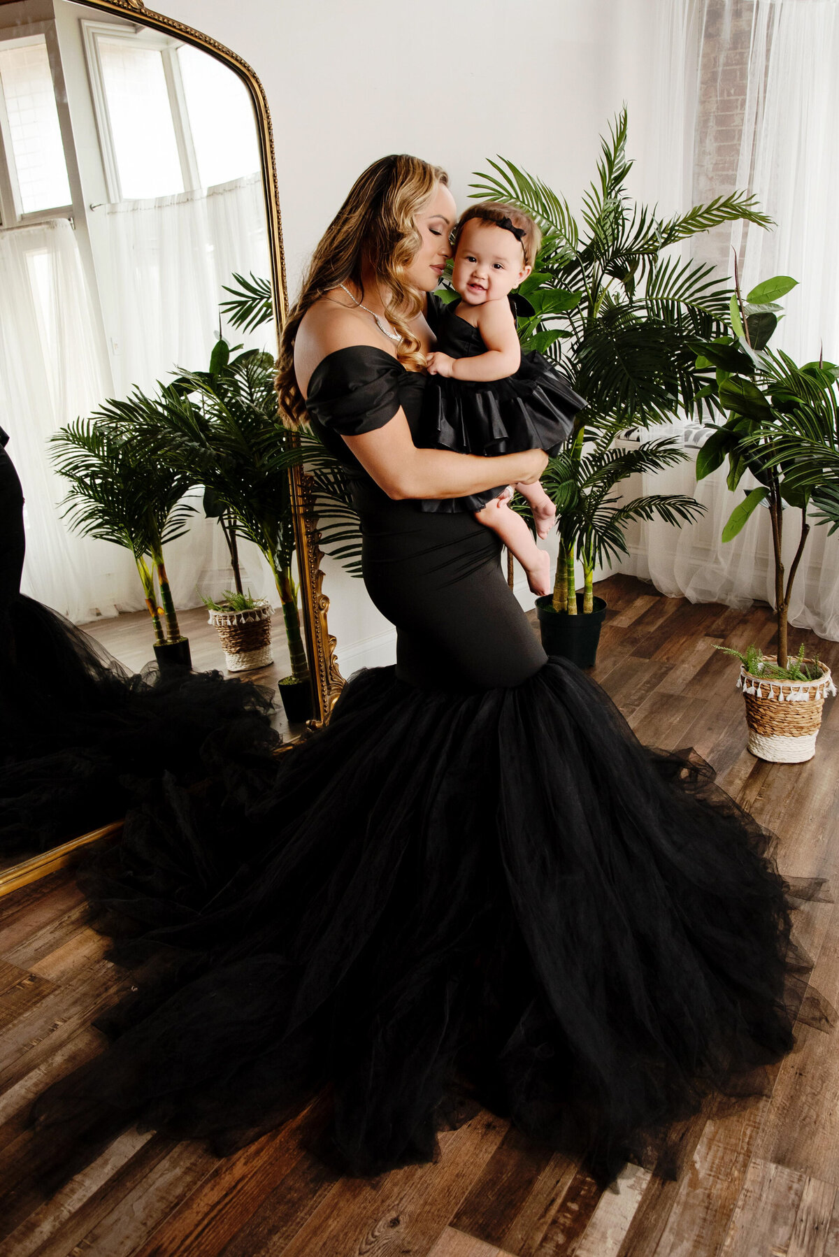 st-louis-family-photographer-mom-in-black-mermaid-gown-with-tulle-bottom-holding-baby-girl-wearing-black-dress-in-front-of-large-mirror-and-plants