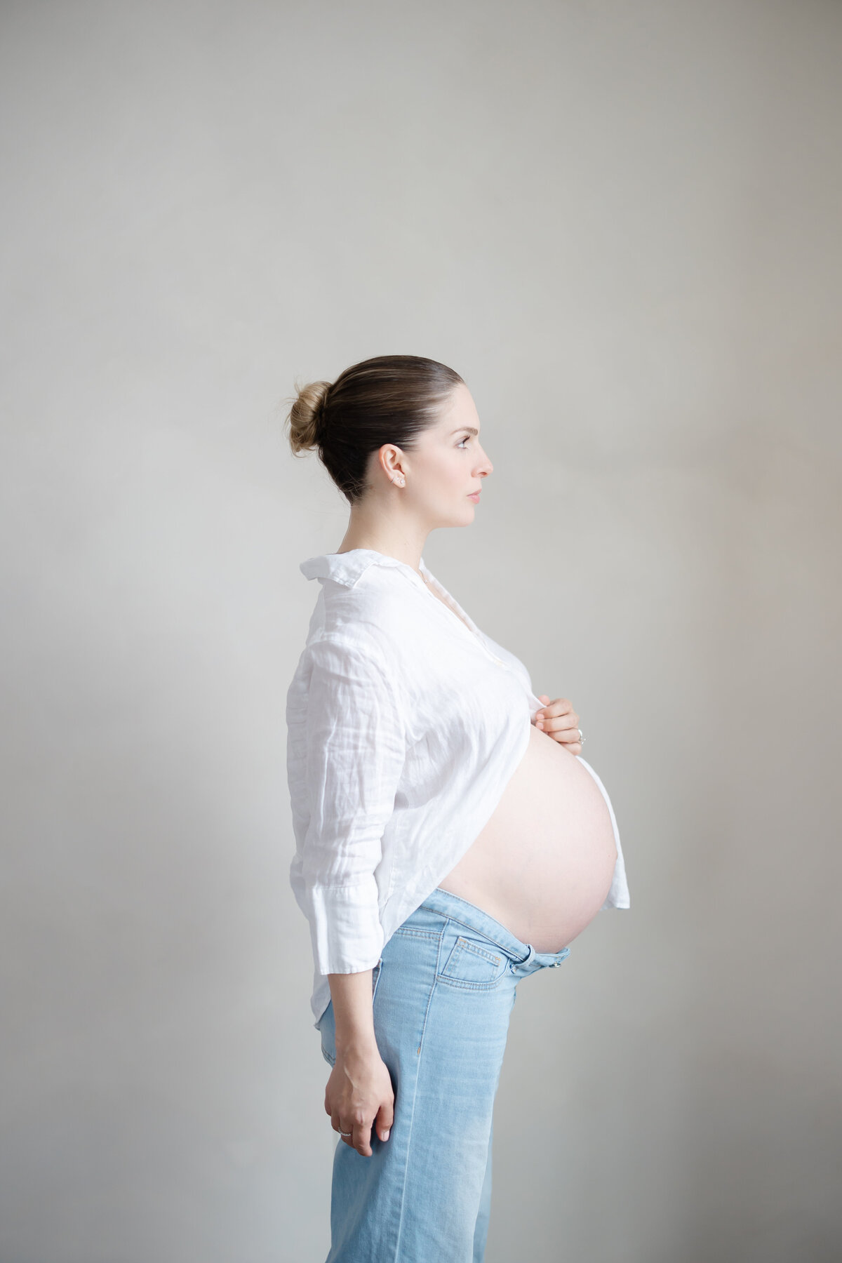 a mother stands in front of a beige limewash wall as her white button up shirt parts for her pregnant belly. She stands to the side, creating a maternity profile pose
