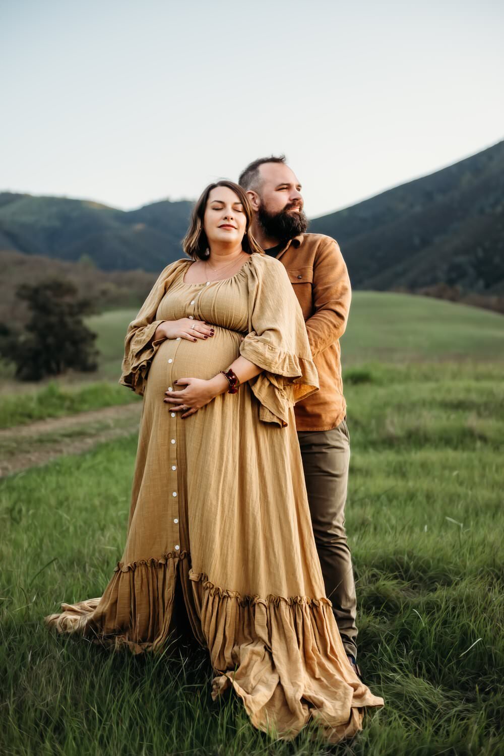 mama and her husband stand at the base of mt diablo. she wears a mustard colored maternity dress.