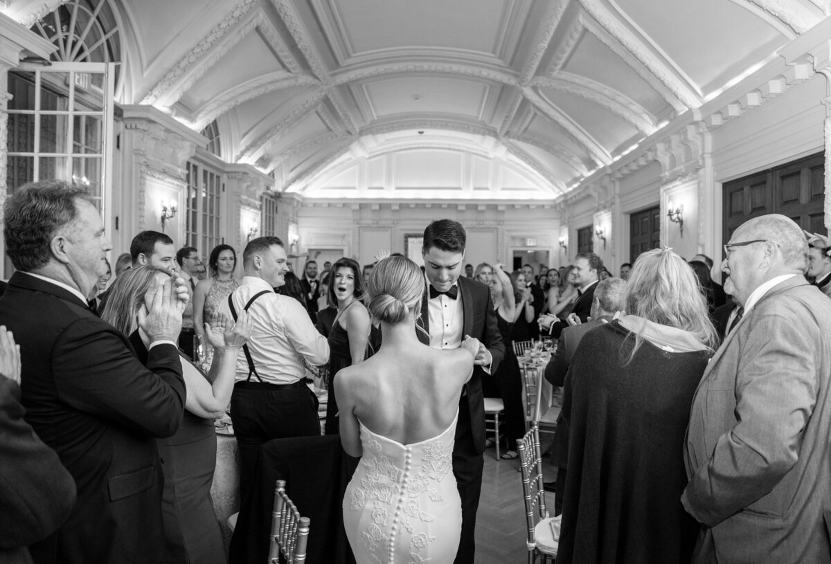 A black and white photo of a wedding reception. The couple stands in the center, dressed in formal attire, as guests surround them, clapping and smiling in a decorated venue with a vaulted ceiling and elegant lighting.