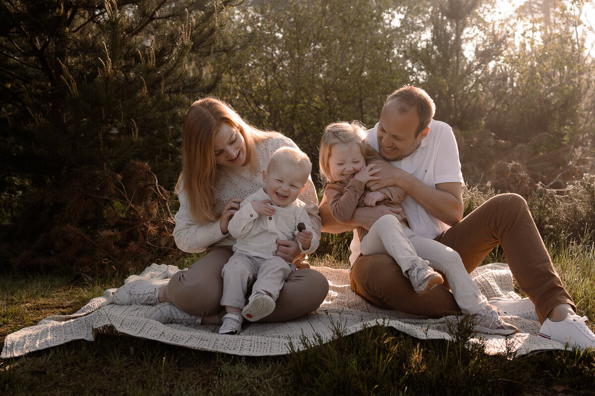Gezin met twee kinderen zitten op het kleedje en zijn elkaar aan het kietelen tijdens een gezinsshoot