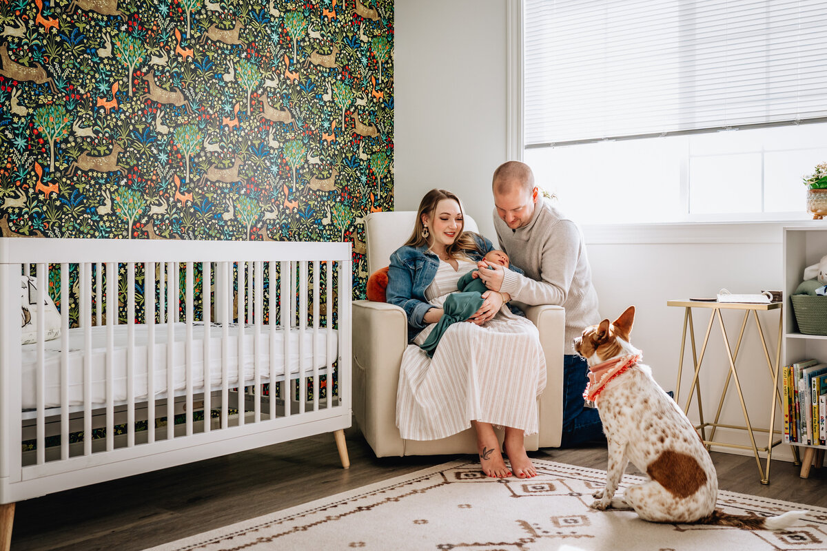 A family of three sits together in a nursery with a colorful woodland-themed wallpaper, their dog sitting attentively beside them, creating a warm and cozy atmosphere.