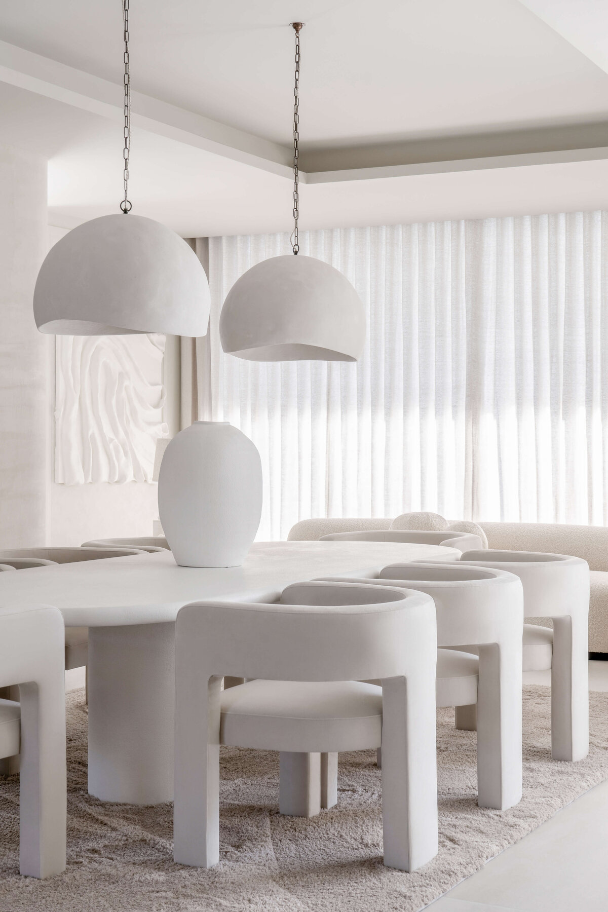 A modern dining room with a minimalist aesthetic, featuring a large oval table surrounded by six curved white chairs. Two large, white dome-shaped pendant lights hang above the table. The room is decorated in neutral tones with a plush, light-colored rug underneath the table and chairs. Behind the table a large white textured art piece can be seen hanging on the wall, adding a contemporary flair to the space.