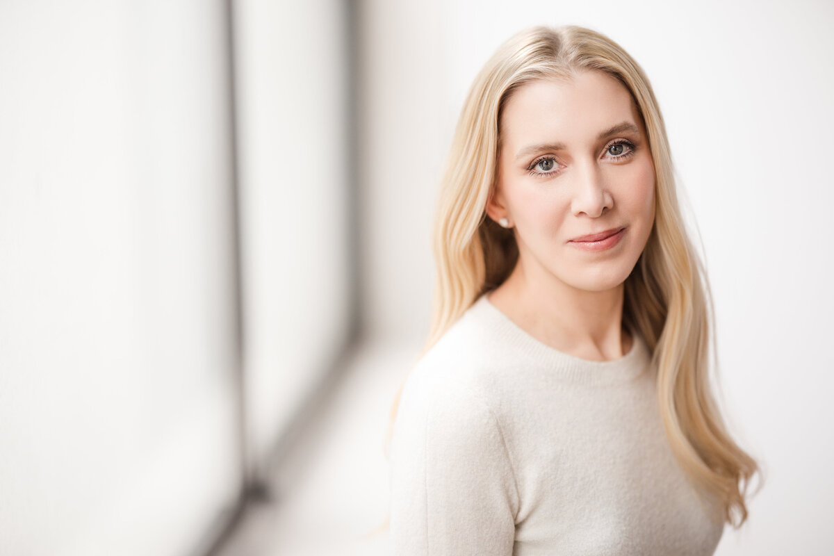 Person with long blonde hair, wearing a light sweater, seated beside a large window with soft natural light. They are looking into the camera with a calm, approachable expression. The background is airy and minimal, creating a bright and clean portrait.
