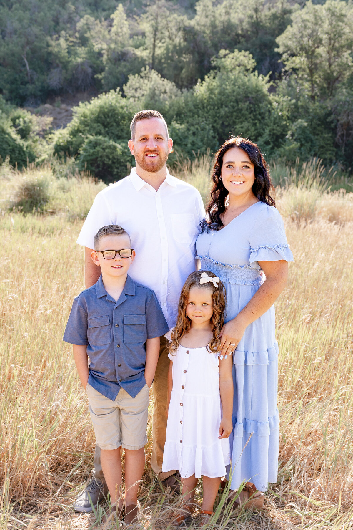 A family of four poses in a sunlit field, with lush green mountains in the background. The father in a white shirt and mother in a light blue dress stand alongside their two children, capturing a timeless family moment.
