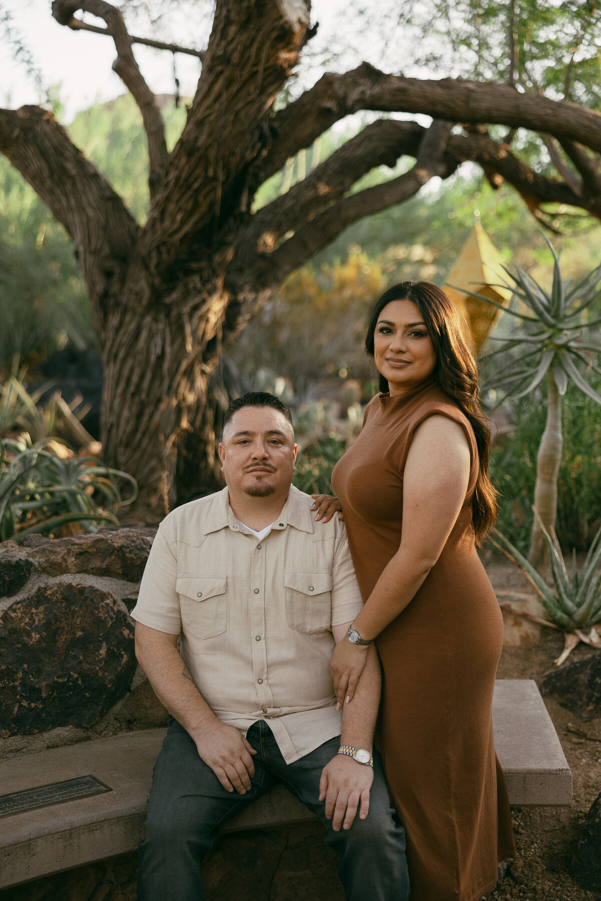 A man in a tan shirt sits on a bench while a woman in a brown dress stands next to him with her hands on his shoulder and arm.