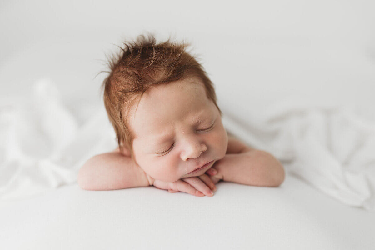 A newborn baby with soft, red hair, resting with arms folded under the chin on a white blanket, eyes closed in a peaceful slumber.