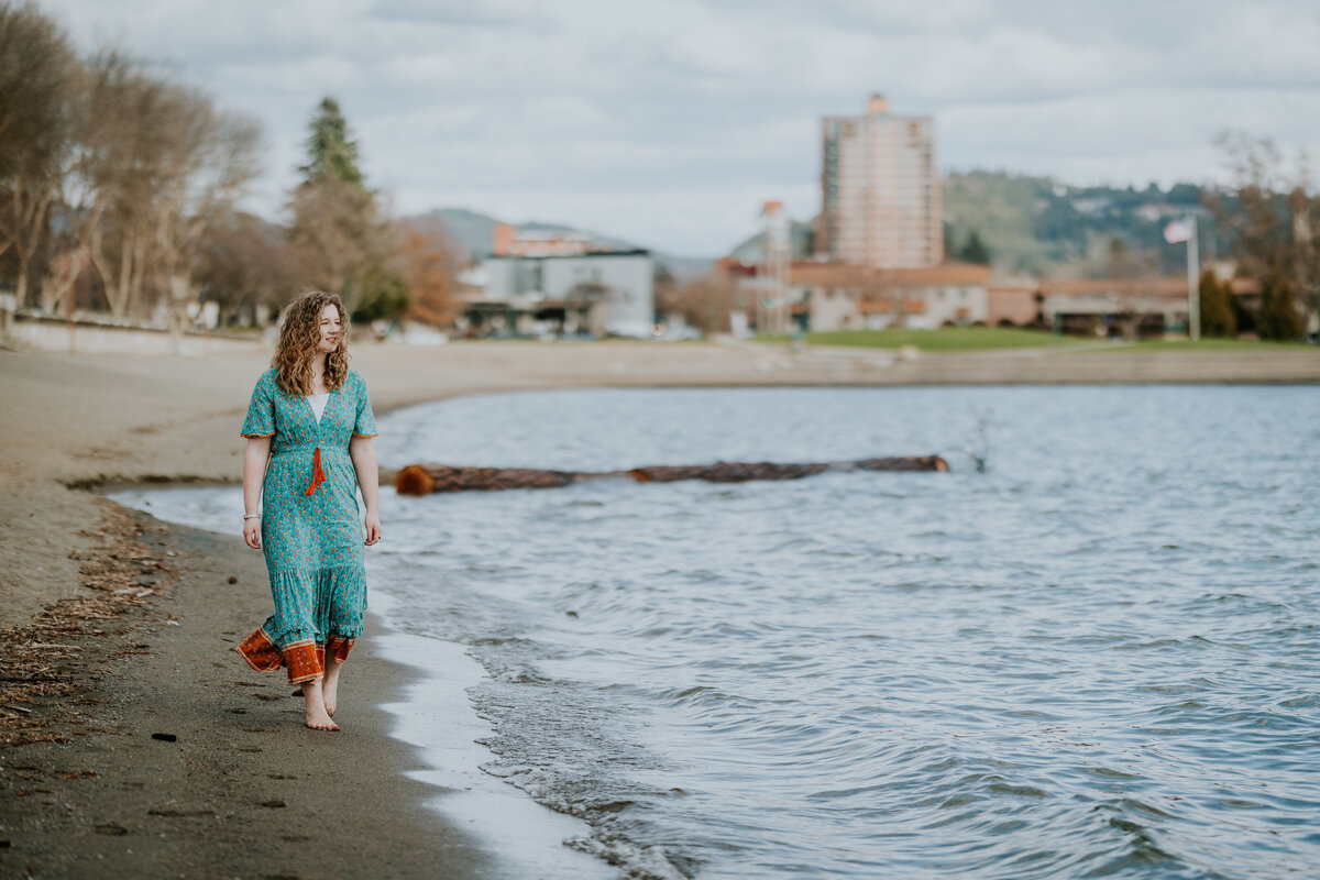 Young woman in teal dress walks along  Coeur d'Alene Lake beach with resort in the background.