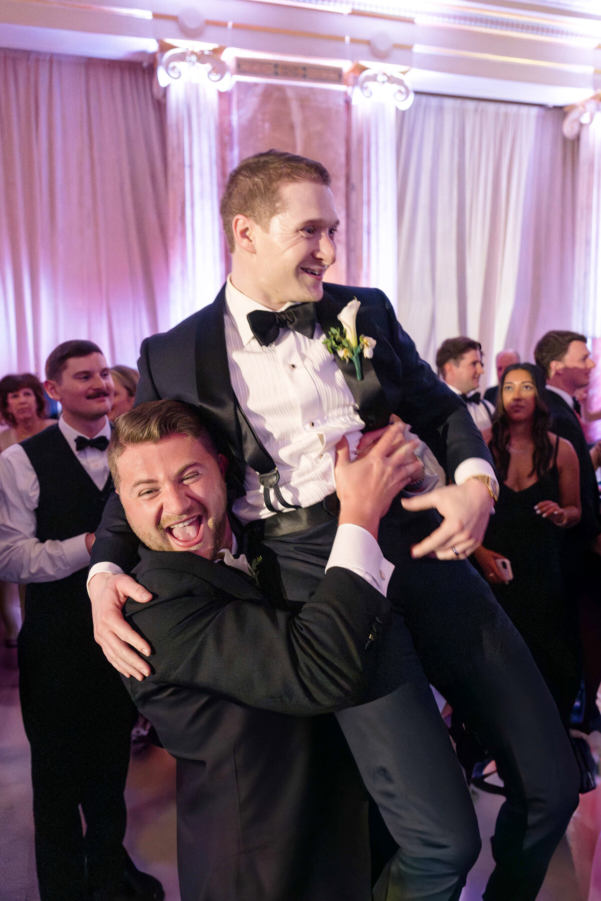 A joyful man in a tuxedo lifts another man in a tuxedo at a lively wedding reception. The background is filled with elegantly dressed guests dancing and celebrating under soft, colorful lighting.