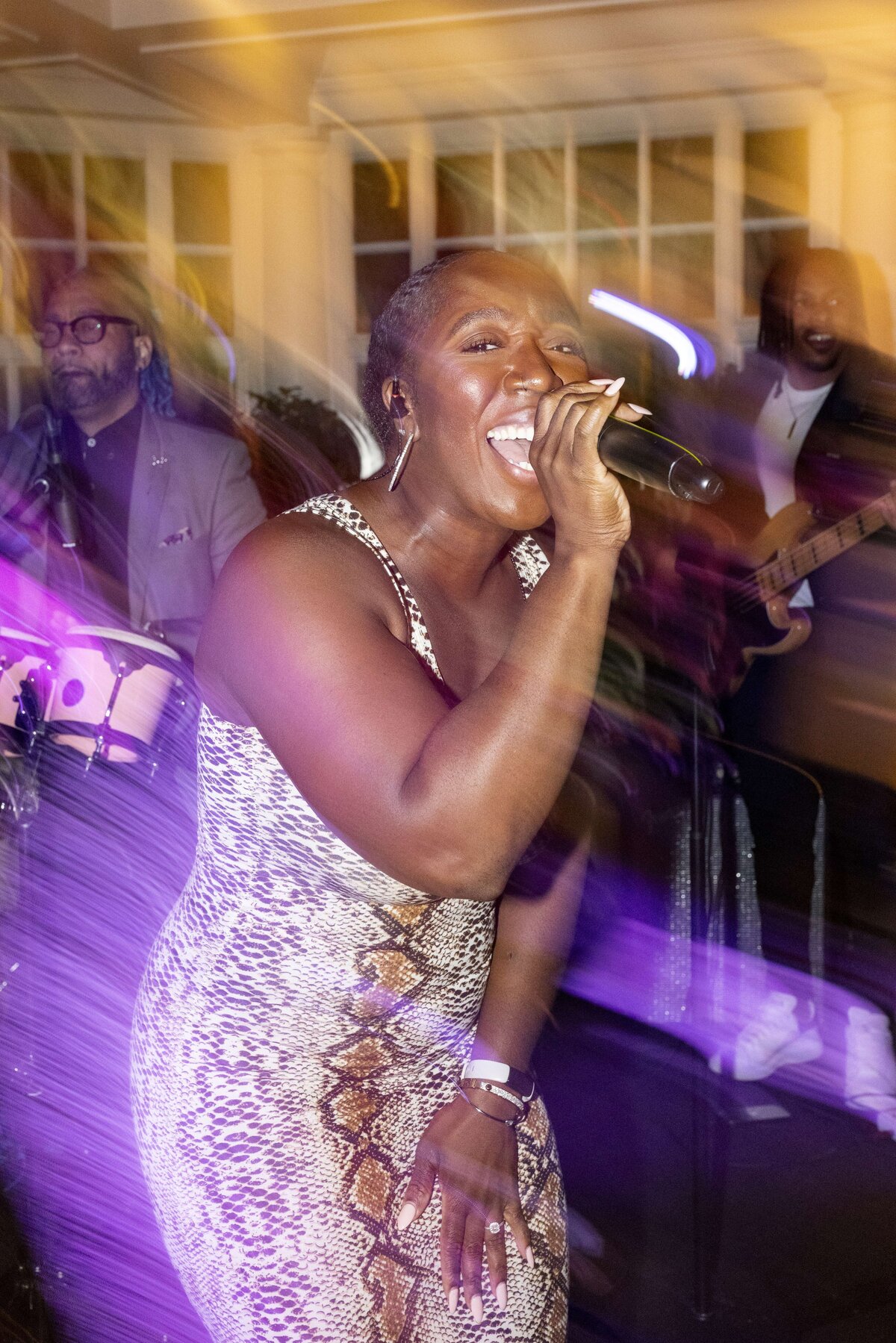 A woman in a patterned dress energetically sings into a microphone. Behind her, musicians play instruments. The scene is colorful and vibrant, with motion blur creating a dynamic effect.
