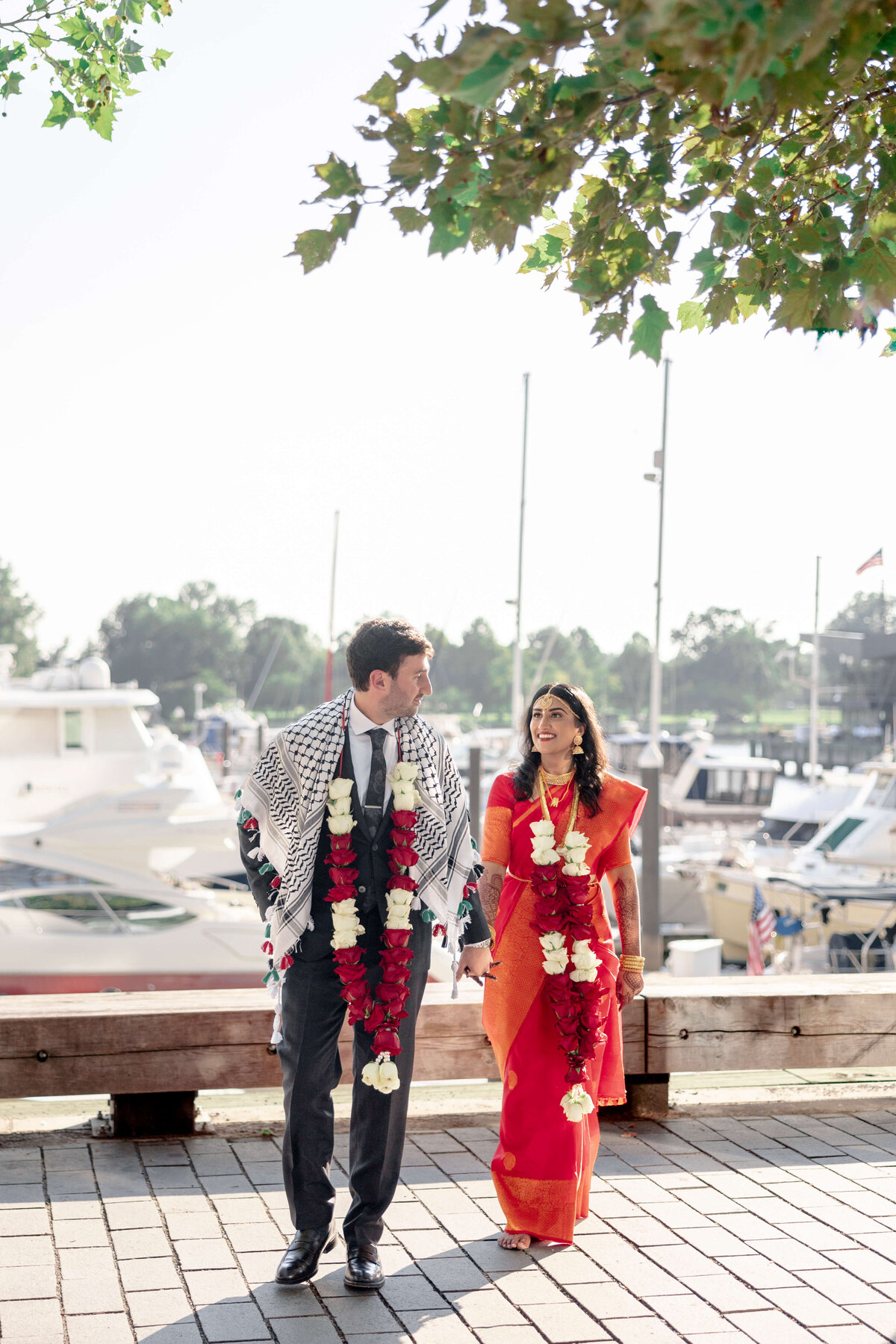 A couple in festive attire walks hand in hand along a waterfront. The man wears a suit with a patterned shawl and garland, and the woman is in a red sari with a garland. Boats and trees are in the background on a sunny day.