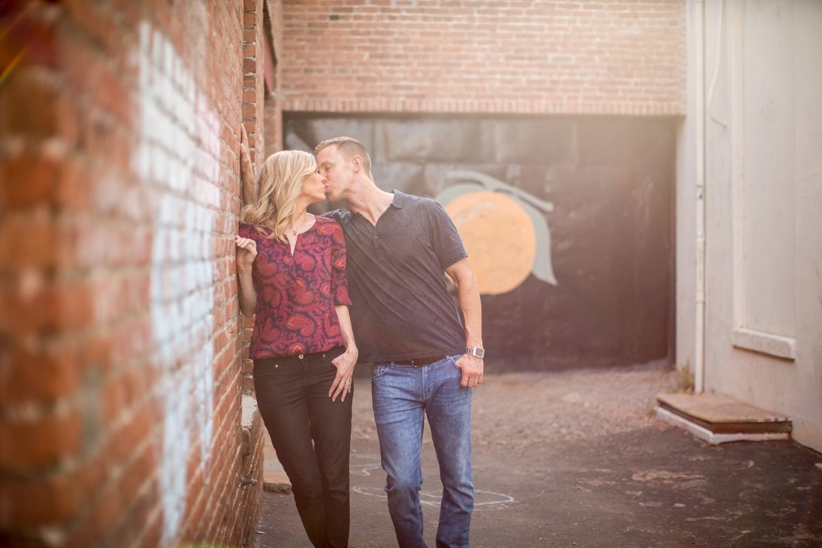 Engaged couple share a kiss during an engagement session along a brick wall in a back alley