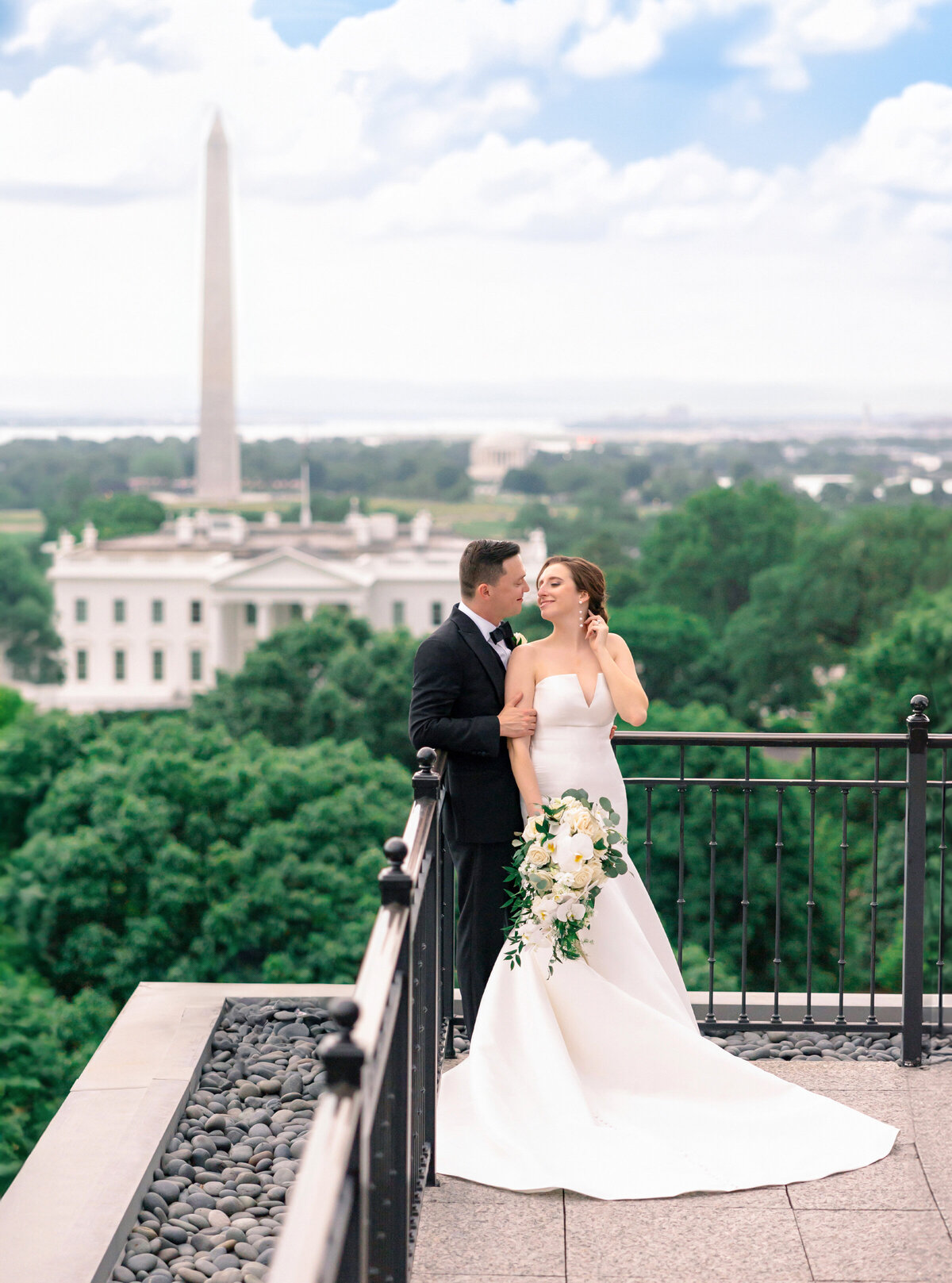 A bride and groom stand on a rooftop with a view of the Washington Monument and the White House in the background. The bride is in a white gown holding a bouquet, and the groom wears a dark suit as they embrace under a blue sky with clouds.