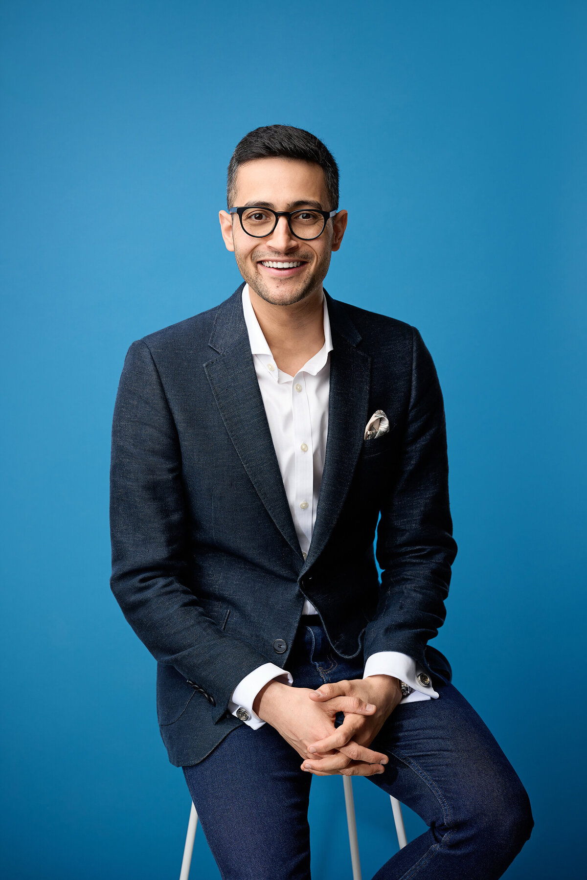 Smiling man on a stool wearing navy jacket and white shirt. He's against a blue background.