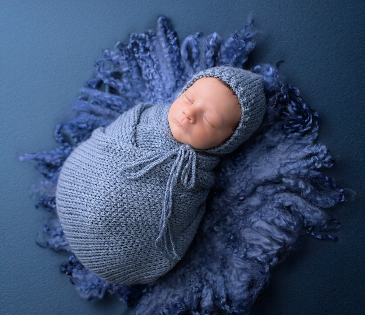 Side angle of a newborn boy wrapped in blue knits with matching hat and placed on similar blue toned textures