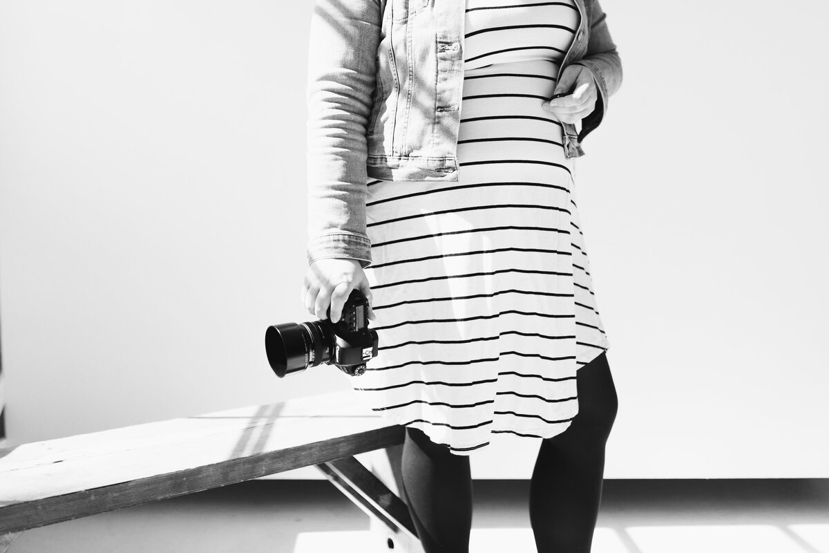photographer stands in studio holding camera, cropped in to just torso and some legs in black and white image