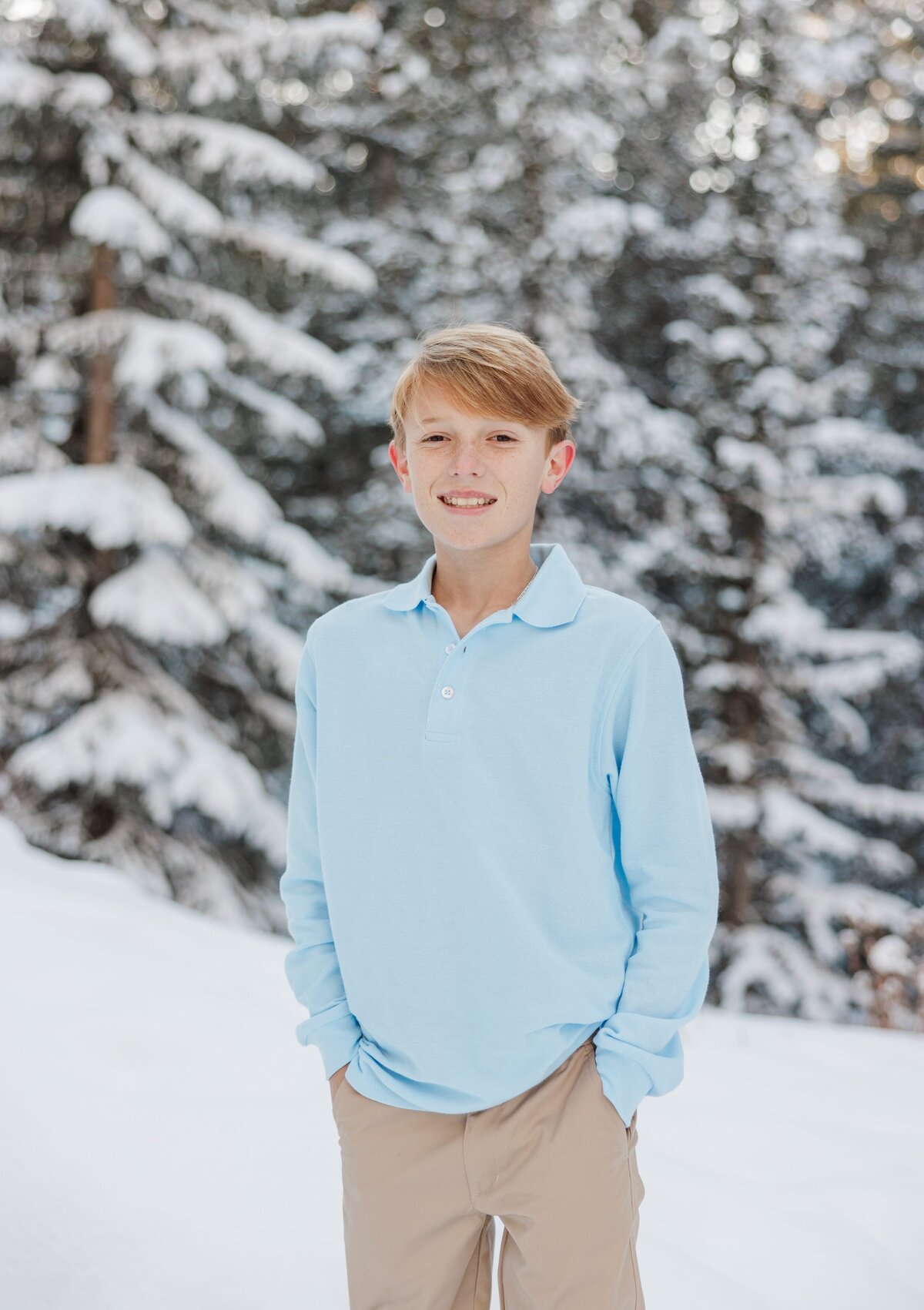 A young boy in a blue polo stands in the snow with hands in pockets