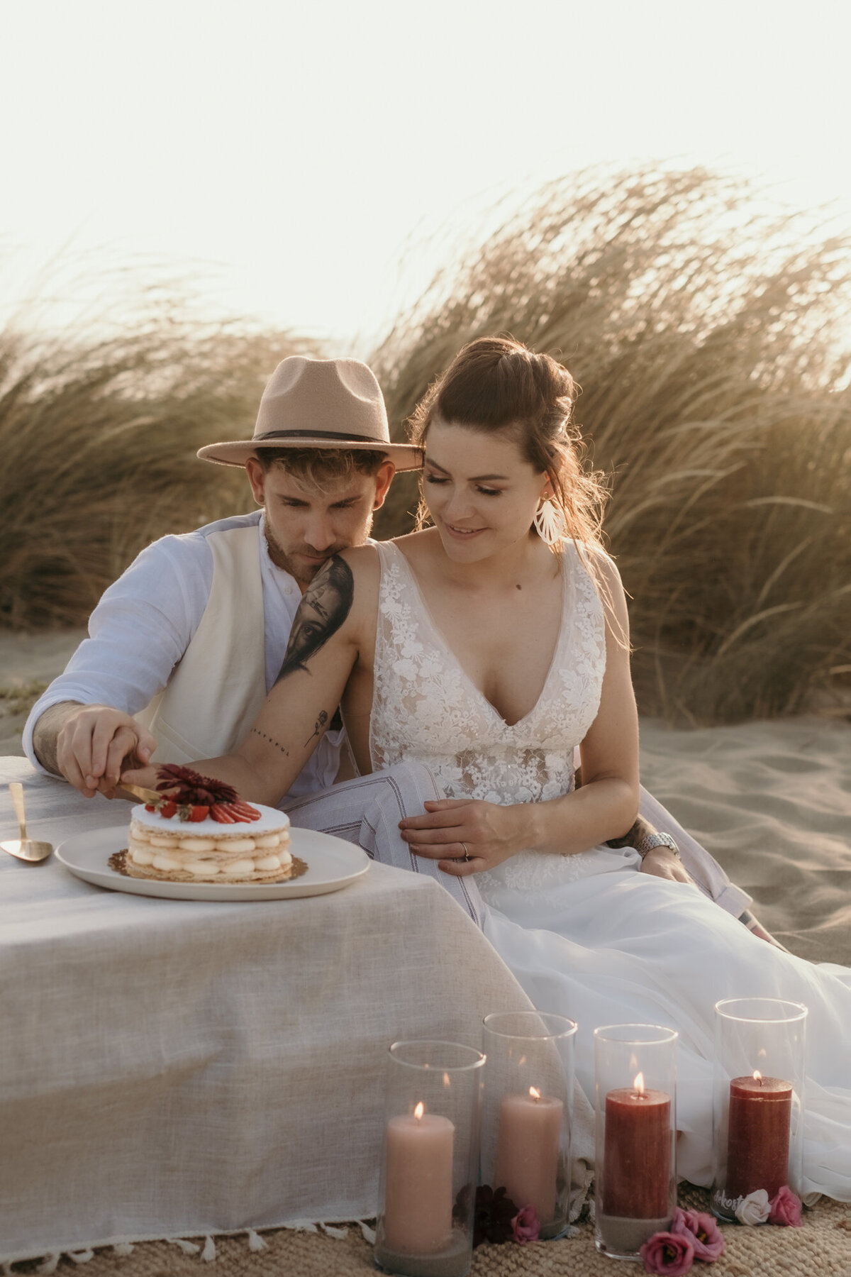 Während des Kuchenanschnitts sitzt das Hochzeitspaar im Sand neben dem niedrigen Tisch hintereinander.