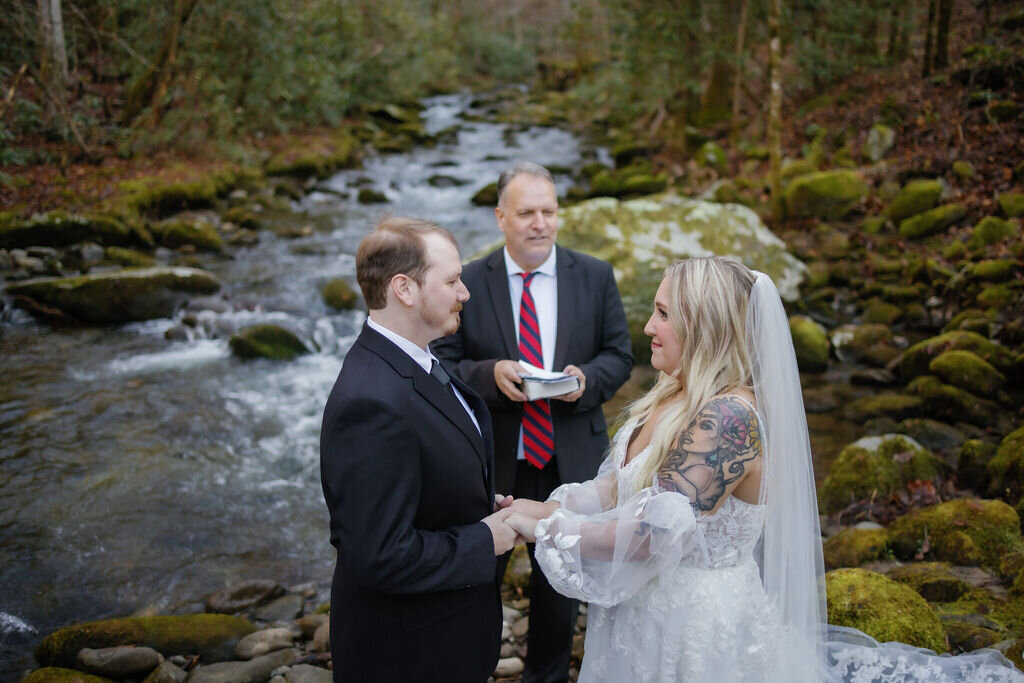gatlinburg photographers captures ceremony photos at Ely's Mill with a river in the background of the woods as the couple eloping to Gatlinburg hold hands and listen to their officiant marry them 